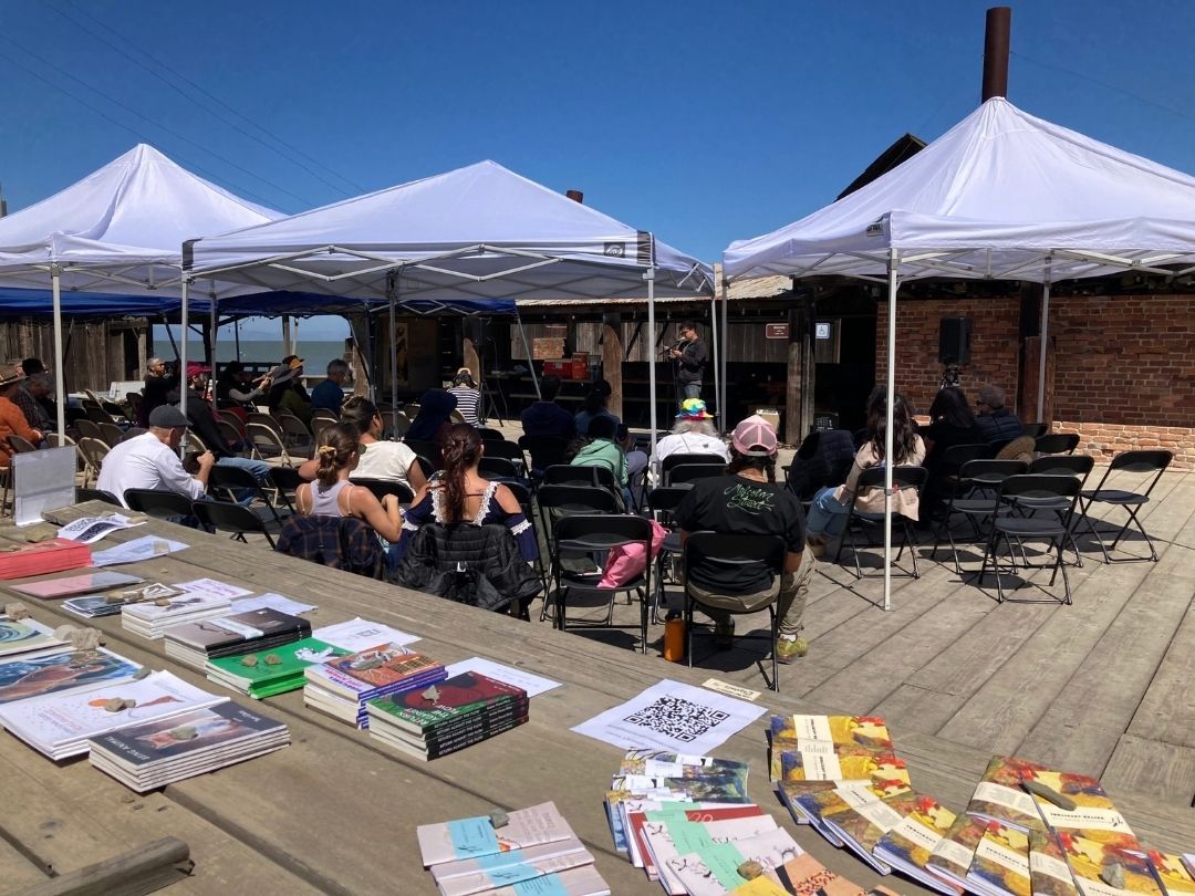 An outdoor event with people seated under white tents, listening to a speaker in front of a brick wall. In the foreground, there is a table displaying various books, pamphlets, and promotional materials.